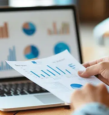 Business analyst reviewing printed revenue growth charts with colorful graphs displayed on laptop screen background